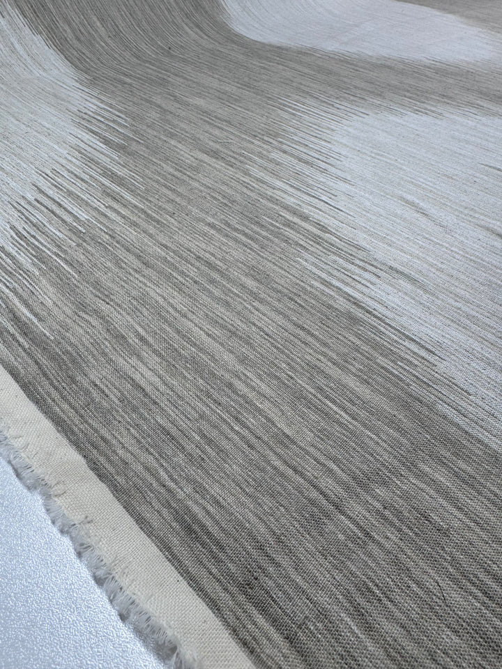The Linen Blend - Solana - 145cm from Super Cheap Fabrics features a gradient of gray and beige creating a flowing, wave-like pattern. Its medium-weight linen presents a sense of movement, while fringed edges add to its textured appearance.