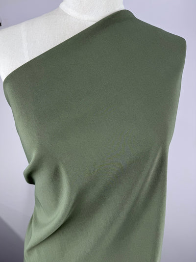 A mannequin gracefully displays the Stretch Bengaline - Beetle (150cm) by Super Cheap Fabrics, styled in an off-shoulder olive-green design. The plain white background enhances the elegance and flow of this spandex blend.