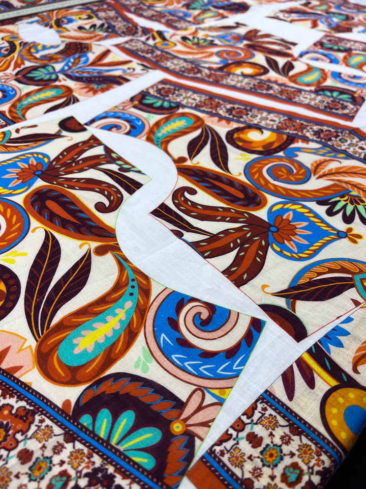 A close-up of Super Cheap Fabrics' 100% Cotton Voile - 130cm panel, featuring vibrant paisley and floral designs in orange, brown, blue, and green with white pattern pieces on top; panel measures 130cm x 140cm.