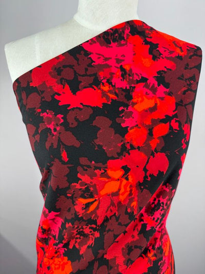 A dress form displays Super Cheap Fabrics' Printed Crepe - Burn - 150cm with a vibrant floral design in red, pink, and black hues. The fabric elegantly drapes over one shoulder, creating a striking contrast against a neutral background.