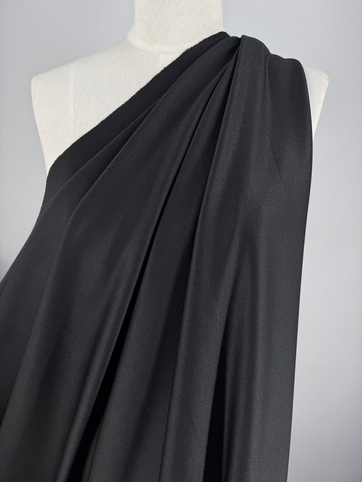 The Textured Knit - Ripple - 155cm by Super Cheap Fabrics, in black polyester, drapes over a mannequin to create soft, flowing folds. The medium weight fabric forms an elegant asymmetrical design against a plain grey background from one shoulder.