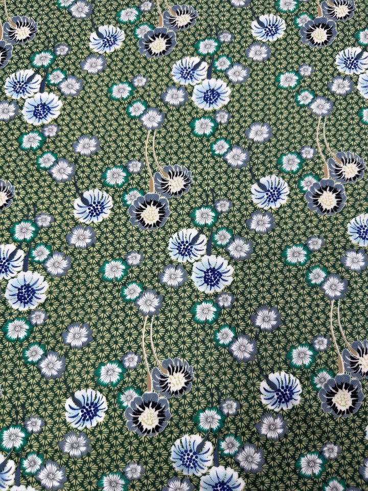 The Printed Crepe - Botan by Super Cheap Fabrics features a green floral pattern with intricate white and blue flowers, accented in dark blue and turquoise. This design includes small leafy details on a 148cm wide crepe fabric.