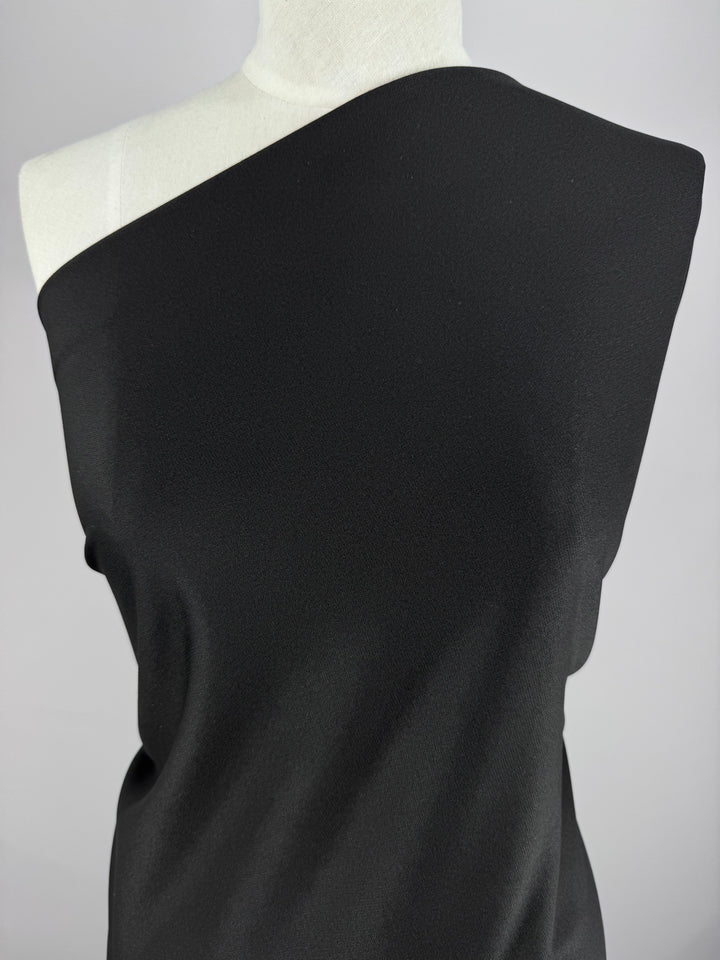 Textured Knit - Ripple - 155cm by Super Cheap Fabrics, a medium-weight black polyester fabric drapes elegantly over a mannequin's shoulder against a light gray backdrop, highlighting its asymmetrical style.