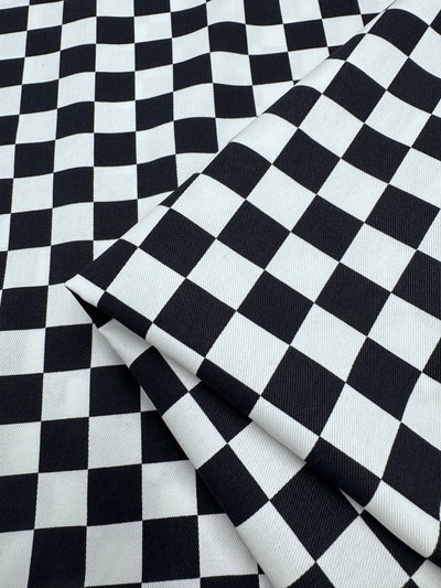 The "Cotton Drill - Checkmate - 148cm" by Super Cheap Fabrics displays a black and white checkered pattern with a folded section in the lower right corner, highlighting its lightweight texture that enhances the repeating squares.