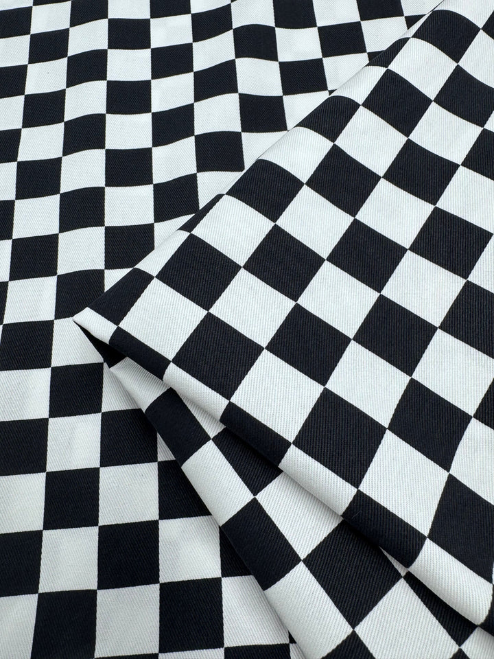 The "Cotton Drill - Checkmate - 148cm" by Super Cheap Fabrics displays a black and white checkered pattern with a folded section in the lower right corner, highlighting its lightweight texture that enhances the repeating squares.