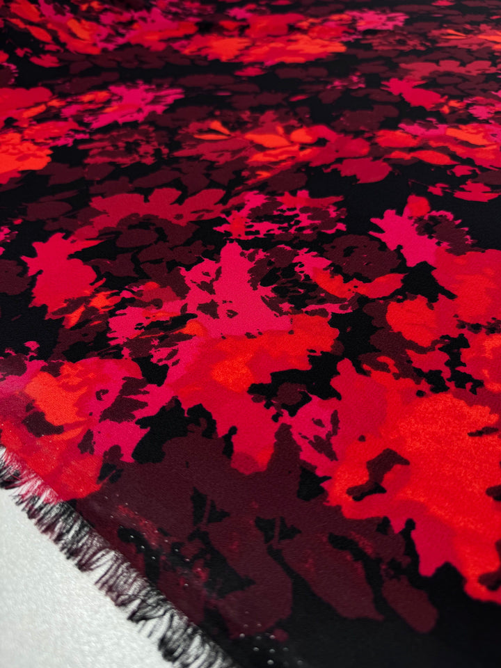 A close-up of "Printed Crepe - Burn - 150cm" by Super Cheap Fabrics shows vibrant crepe with an abstract floral design in red, orange, and pink on black. The slightly frayed edges add texture, ideal for bold fashion garments.