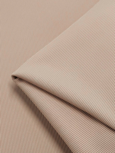 The Rib Knit - Biscuit by Super Cheap Fabrics features folded beige fabric with thin, parallel white stripes forming a pinstripe pattern. The folds converge at the bottom right corner, enhancing its textured surface.