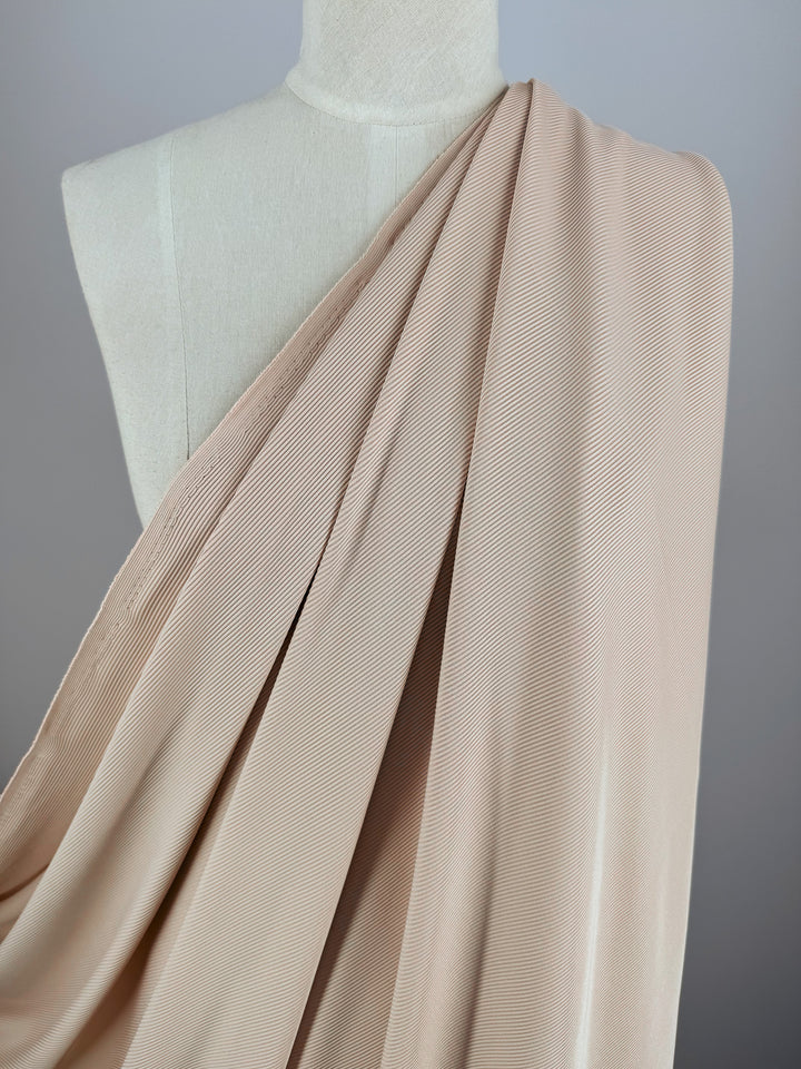 A mannequin features the Rib Knit - Biscuit - 130cm by Super Cheap Fabrics, with its light beige textured knit fabric arranged diagonally, showcasing gentle folds against a neutral backdrop for an elegant look.