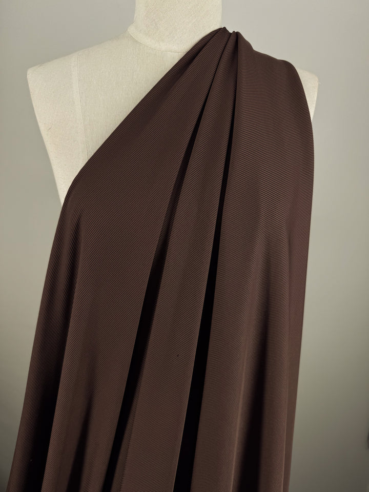 A mannequin is elegantly draped in Super Cheap Fabrics' Rib Knit - Truffle - 130cm, a rich brown textured knit. This medium-weight fabric hangs in pleats over one shoulder like a toga against a light gray background.
