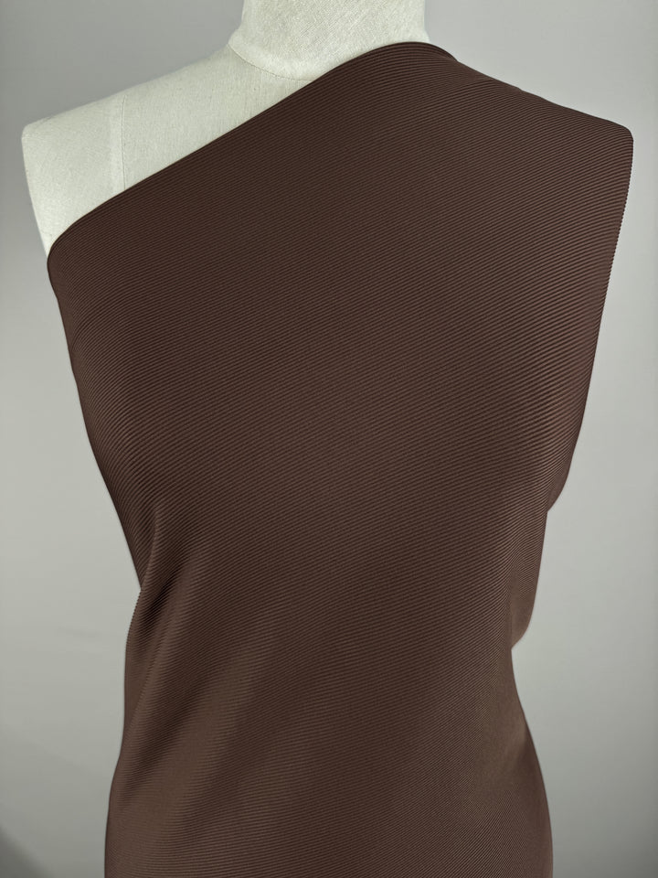 A mannequin displays a medium-weight Rib Knit - Truffle (130cm) garment by Super Cheap Fabrics, featuring a one-shoulder design in brown. The plain light gray background highlights the textured knit fabric beautifully.