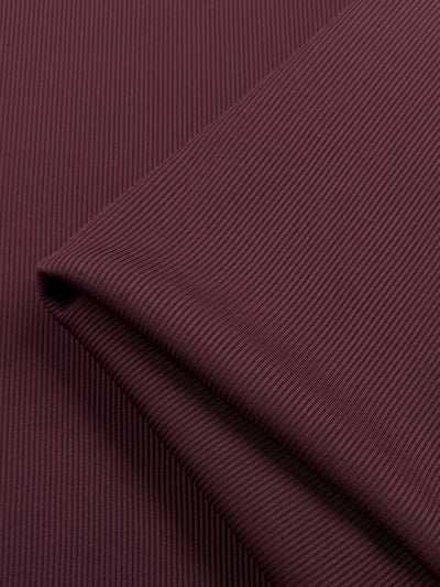 A close-up showcases the Rib Knit - Cherry fabric by Super Cheap Fabrics. This dark maroon, medium-weight fabric features a fine vertical texture and stretch, folded at the top right corner, highlighting a smooth play of light and shadow across its surface. Width: 130cm.