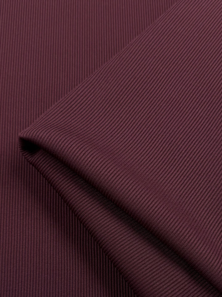 A close-up showcases the Rib Knit - Cherry fabric by Super Cheap Fabrics. This dark maroon, medium-weight fabric features a fine vertical texture and stretch, folded at the top right corner, highlighting a smooth play of light and shadow across its surface. Width: 130cm.