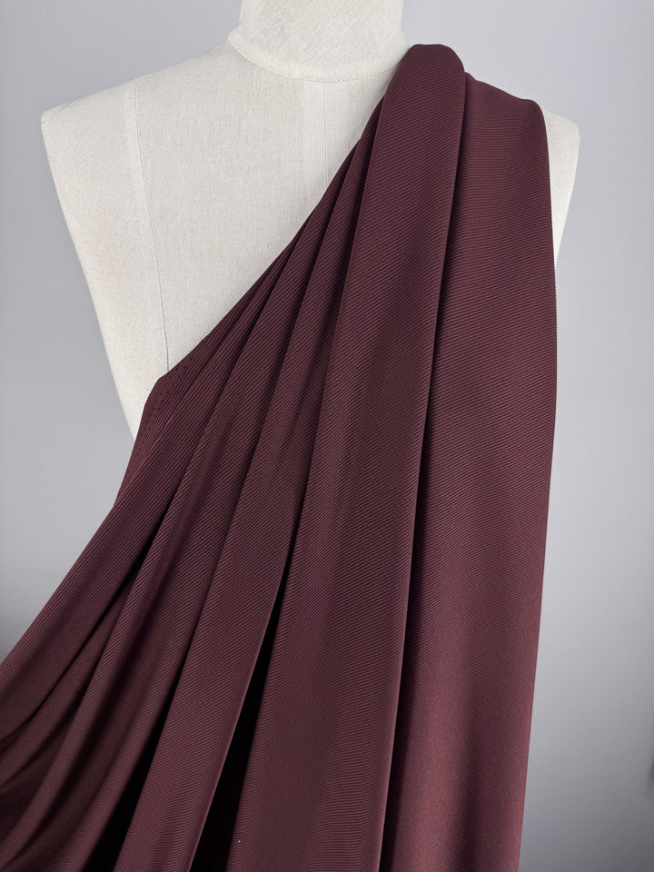 The Rib Knit - Cherry - 130cm fabric from Super Cheap Fabrics drapes elegantly on a dress form, featuring medium weight stretch in textured burgundy over a plain gray backdrop, accentuating its rich color and smooth folds.