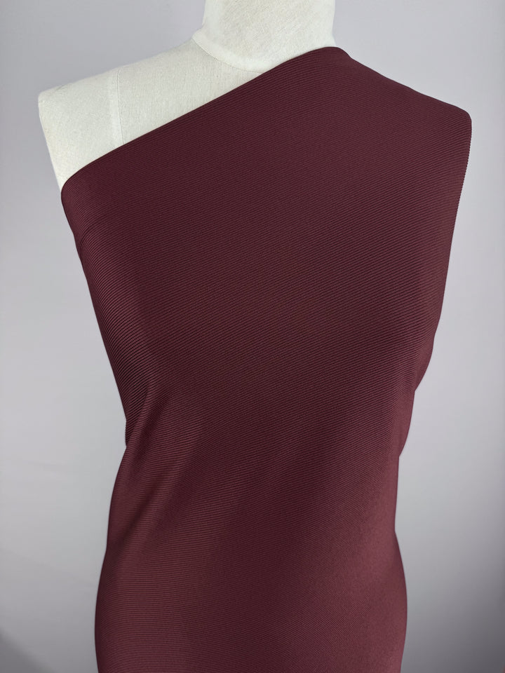 A mannequin elegantly wears the "Rib Knit - Cherry - 130cm" by Super Cheap Fabrics, a one-shoulder burgundy garment crafted from medium weight rib knit, set against a light gray background.