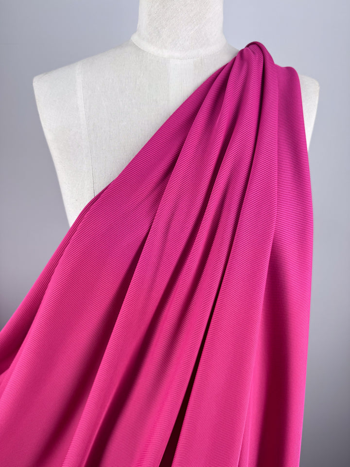 A mannequin displays the Rib Knit in Hot Pink (130cm) by Super Cheap Fabrics, arranged in gentle folds against a gray backdrop, highlighting the allure of textured knit fabrics.