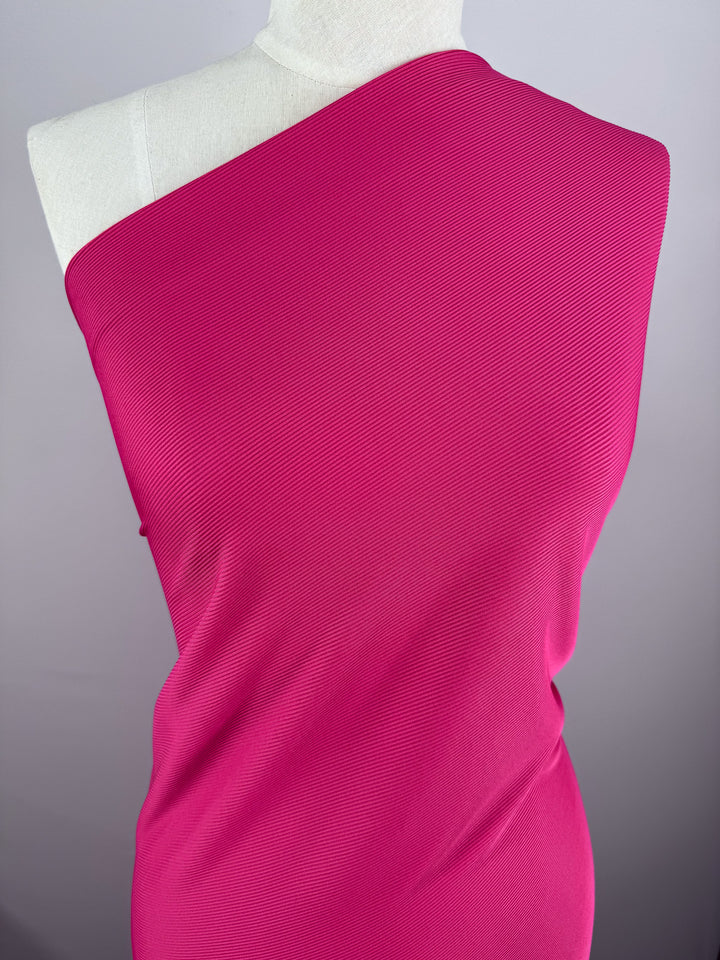 A mannequin showcases the "Rib Knit - Hot Pink - 130cm" fabric by Super Cheap Fabrics, draped stylishly over one shoulder. The dynamic rib-knit pattern offers a modern look, set against a plain gray background.