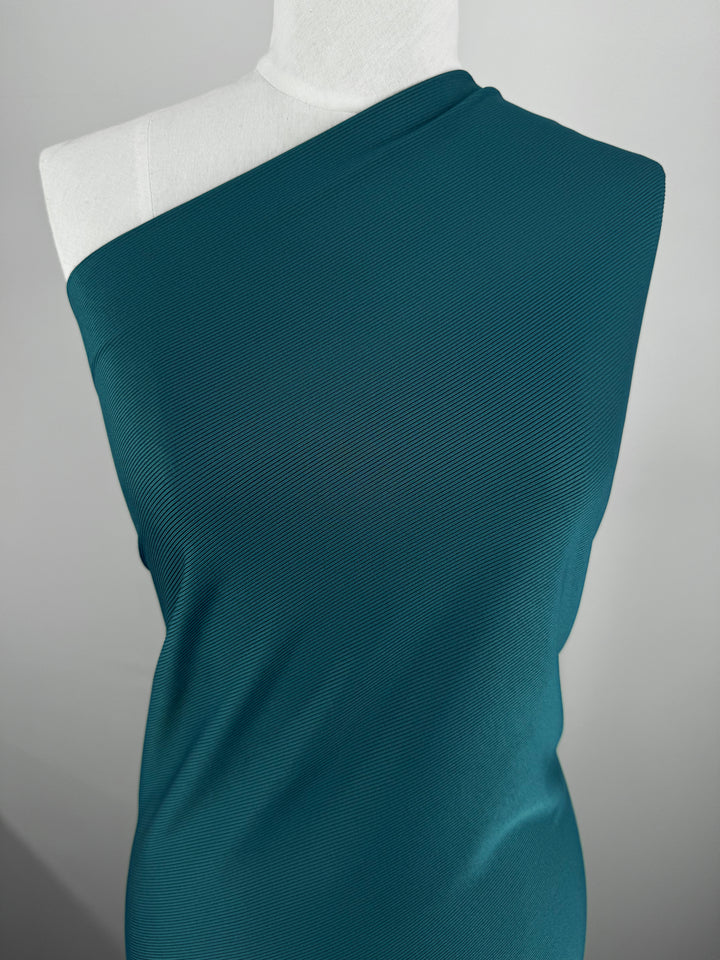 A mannequin displays the 'Rib Knit 1x1 - Teal - 125cm' by Super Cheap Fabrics, a textured teal one-shoulder garment. The medium-weight rib knit provides generous stretch, with its soft grey background enhancing the fabric's vibrant color and texture.