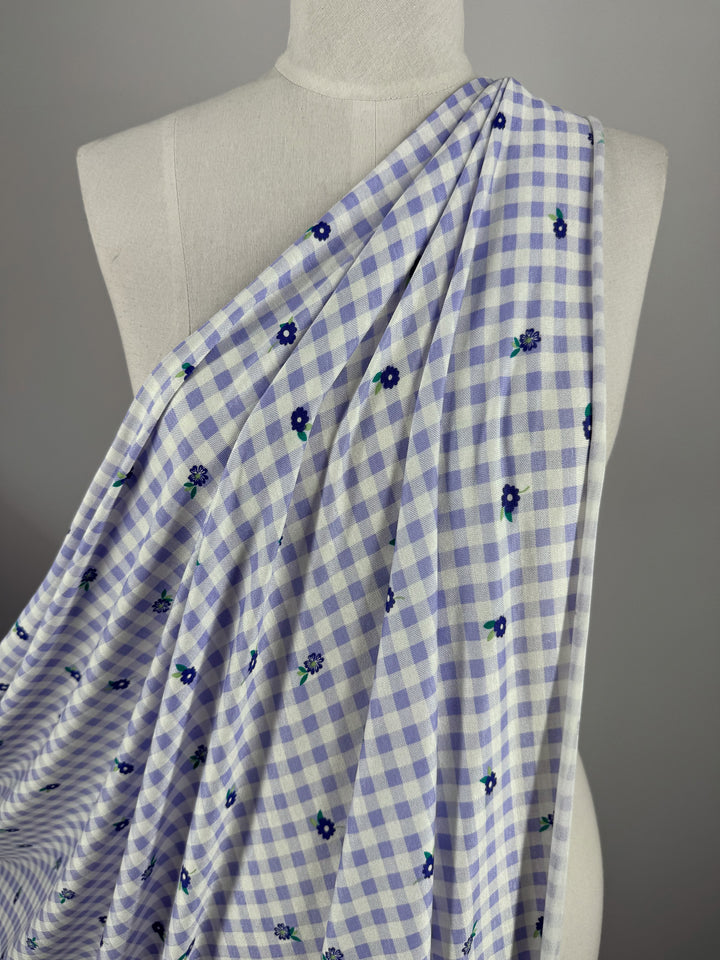 The Printed Cotton Lycra - Lavender Ditsy by Super Cheap Fabrics, 153cm wide, draped over a mannequin. It features a white and lavender checkered pattern with small floral motifs for a delightful touch.