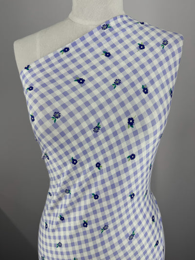 A close-up of a mannequin draped in Super Cheap Fabrics' Printed Cotton Lycra - Lavender Ditsy, showcases white and light purple checkered fabric adorned with dark blue flowers, styled asymmetrically across the shoulders for an elegant touch. Width: 153cm.