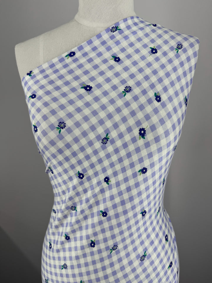 A close-up of a mannequin draped in Super Cheap Fabrics' Printed Cotton Lycra - Lavender Ditsy, showcases white and light purple checkered fabric adorned with dark blue flowers, styled asymmetrically across the shoulders for an elegant touch. Width: 153cm.