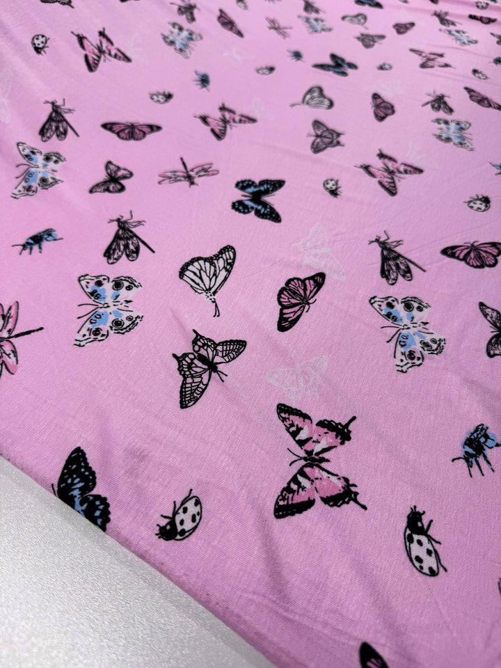 The "Printed Cotton Lycra - Pink Fly - 150cm" from Super Cheap Fabrics combines pink cotton spandex with black and blue butterflies, moths, and ladybugs. Its light to medium weight allows it to drape beautifully for a whimsical, colorful look.