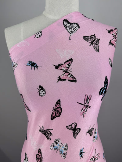 The Super Cheap Fabrics' Printed Cotton Lycra - Pink Fly, measuring 150cm, is displayed on a dress form. Its pink fabric with black and white butterfly and beetle prints offers both comfort and style due to its light-medium weight cotton spandex blend, enhancing the intricate pattern against a neutral backdrop.