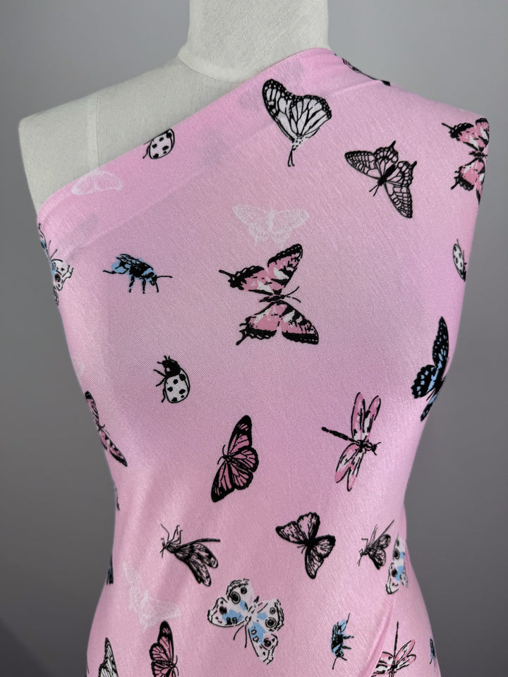 The Super Cheap Fabrics' Printed Cotton Lycra - Pink Fly, measuring 150cm, is displayed on a dress form. Its pink fabric with black and white butterfly and beetle prints offers both comfort and style due to its light-medium weight cotton spandex blend, enhancing the intricate pattern against a neutral backdrop.