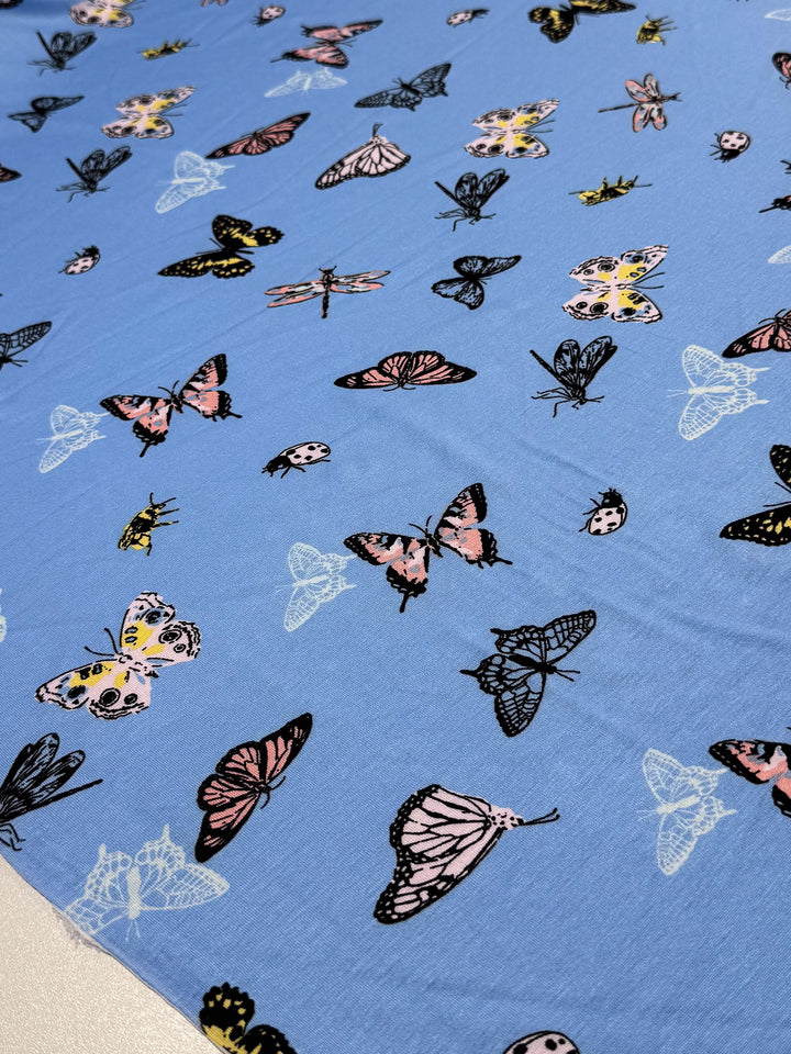 The Super Cheap Fabrics Printed Cotton Lycra - Blue Butter, 155cm features a vibrant design of butterflies and moths in pink, black, white, and yellow.