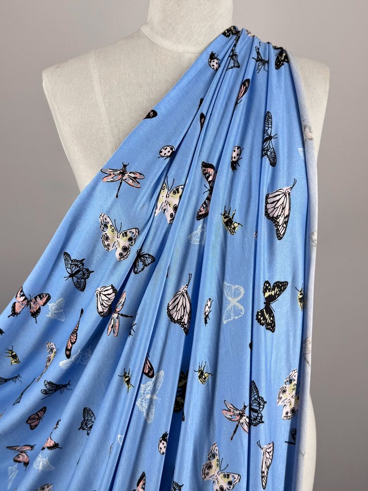 The 155cm Printed Cotton Lycra - Blue Butter from Super Cheap Fabrics is displayed over a mannequin, featuring black, pink, and white butterfly and insect designs on a light blue background with a solid light gray backdrop for an elegant contrast.
