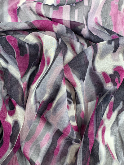 A close-up of crumpled, elegant fabric with an abstract pattern in shades of pink, gray, white, and black. The flowing design has brushstroke lines and a textured look similar to Super Cheap Fabrics' Pure Silk - Electric Mirage - 132cm—ideal for high-end fashion with its dynamic and soft effect.