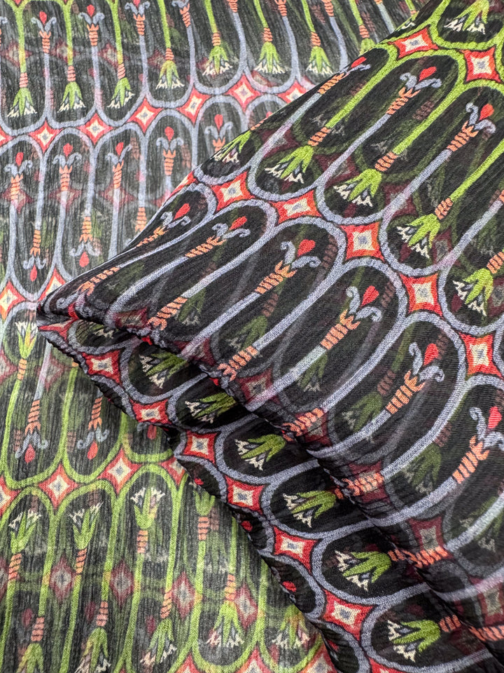 This luxury fabric from Super Cheap Fabrics, named Pure Silk - Serpent Bloom - 135cm, features colorful abstract patterns with vertical leaf-like designs in green, red, and black. The motifs are enclosed in oval shapes with red diamond accents and the silk is folded to reveal its elegant layers.