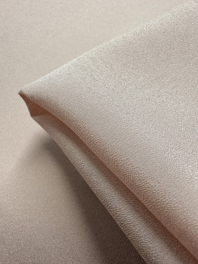 Close-up of folded mauve chalk crepe fabric from Super Cheap Fabrics, 150cm wide, with a light beige hue and subtle sheen. Smooth, soft texture enhanced by gentle lighting emphasizing its silky surface.