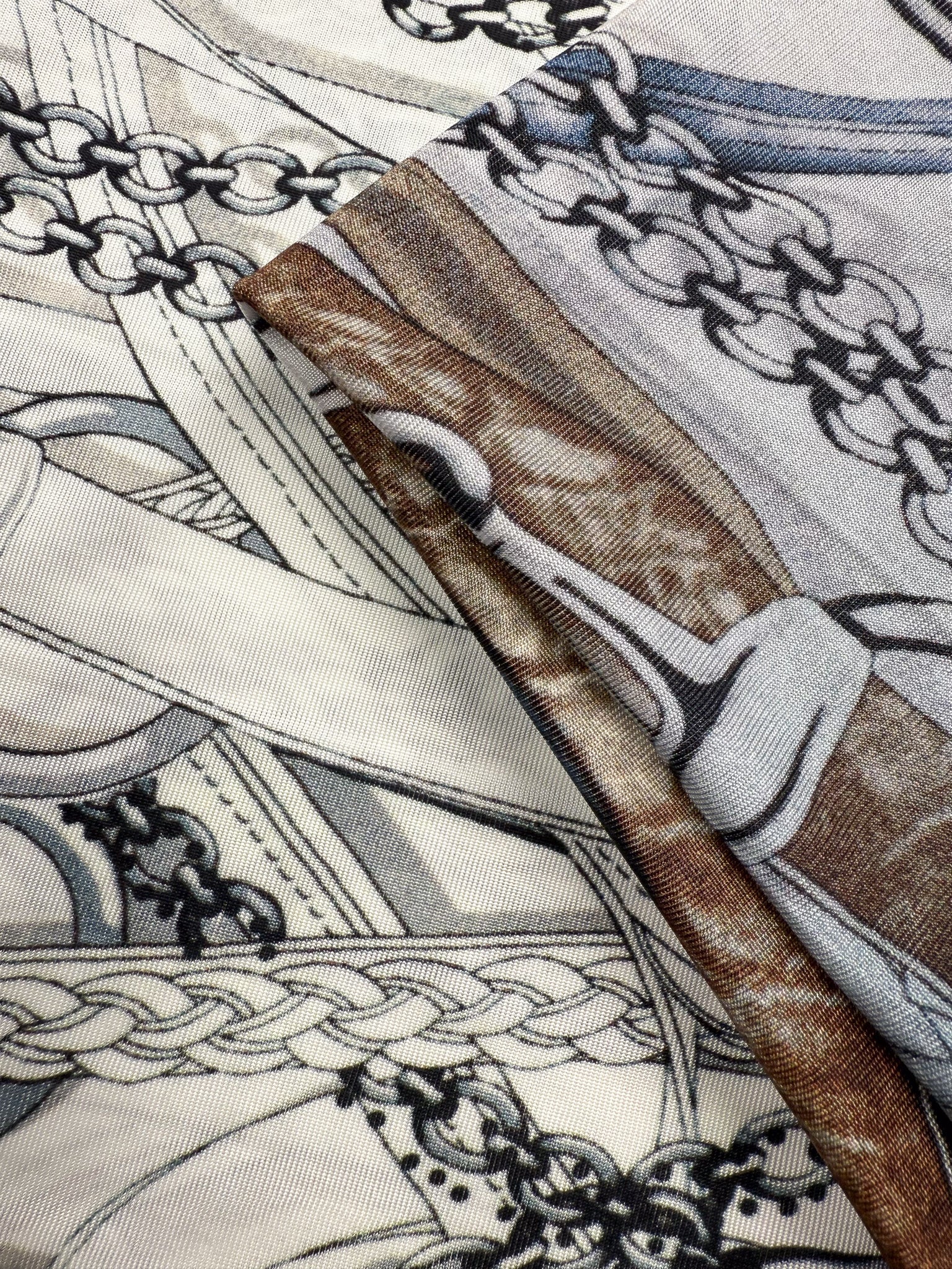Close-up of Super Cheap Fabrics' Viscose Knit - Link (138cm), showcasing two pieces with intricate equestrian-themed patterns—interwoven chains, ropes, and stirrups in cream, gray, black, and brown.