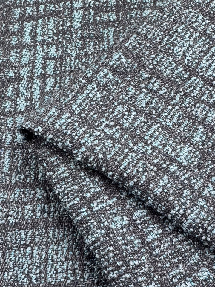 A close-up of Super Cheap Fabrics' Upholstery - Dexter 37 - 147cm shows a gray and light blue woven pattern, made from polyester and viscose. The fabric's folded thickness highlights a grid-like design with a speckled appearance.