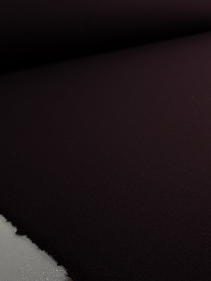 A close-up of Super Cheap Fabrics' Wool Cashmere - Gothic Grape (150cm) shows the soft, smooth maroon fabric unrolled flat with frayed edges at the bottom left, highlighting its luxurious texture.