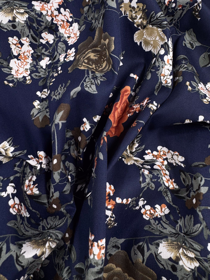 A close-up of Super Cheap Fabrics' Cotton Sateen - Autumn Romance (145cm), showing its navy background with orange, white, and olive green floral print. This lightweight, slightly wrinkled cotton is ideal for summer dresses.
