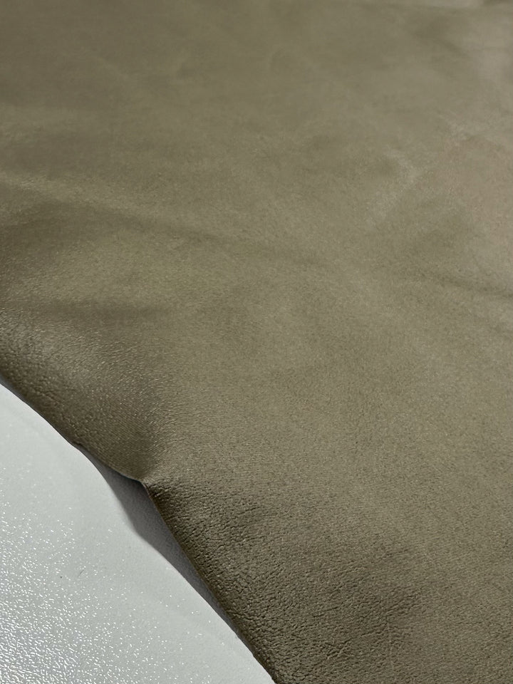 A close-up of Super Cheap Fabrics' Faux Spandex Leather - Tea Leaf, shows its smooth, light brown texture reminiscent of high-end fabric. Laid over a glossy white surface, it creates subtle shadows along the edges.
