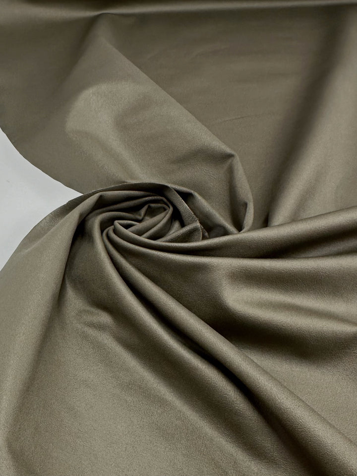 A close-up of Tea Leaf Faux Spandex Leather from Super Cheap Fabrics, elegantly draped in an olive hue with a smooth, shiny texture. This high-end polyester spandex fabric is gathered in a swirl pattern, creating fluid curves and folds. Width: 145cm.