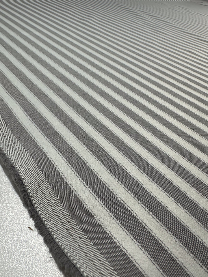 A close-up of Super Cheap Fabrics' Upholstery - Silver & Grey - 140cm shows a silver grey, medium-weight fabric with alternating dark and light gray stripes. The 100% polyester texture is visible against the subtly reflective surface beneath, with slightly frayed edges.