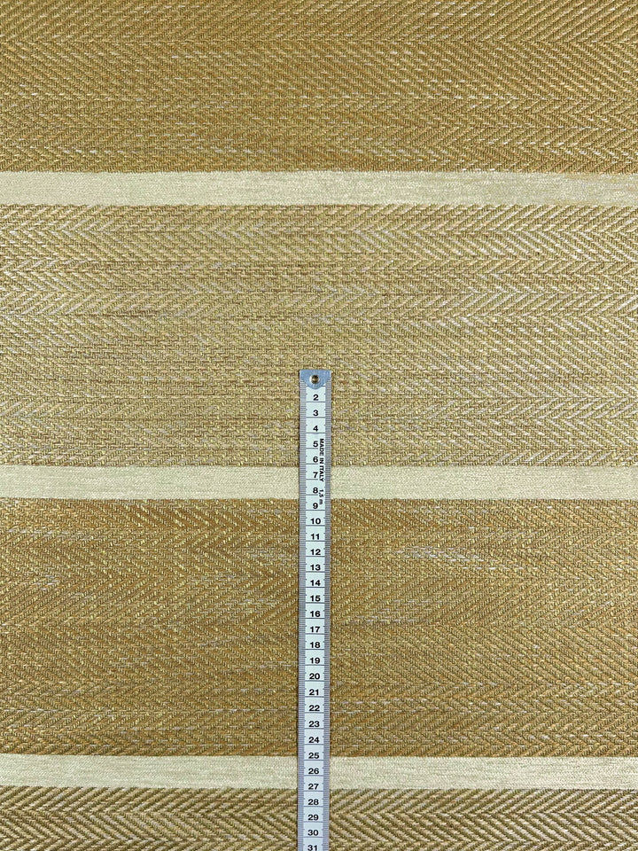 The Upholstery Herringbone - Hickory fabric by Super Cheap Fabrics features a textured, golden hue with a subtle herringbone weave and chevron patterns. It has three horizontal stripes and a vertical measuring tape marked in centimeters up to 24 cm is placed in the center. Fabric width: 148 cm.