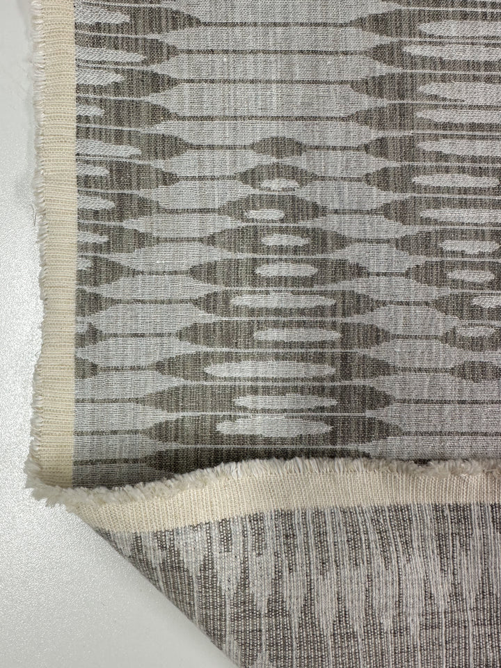 Close-up of Super Cheap Fabrics' Upholstery Jacquard - Ludwig Silver, a textured weave fabric with an abstract geometric pattern in white, gray, and beige shades. It features a fringed edge and is shown layered on a light surface. Width: 140cm.