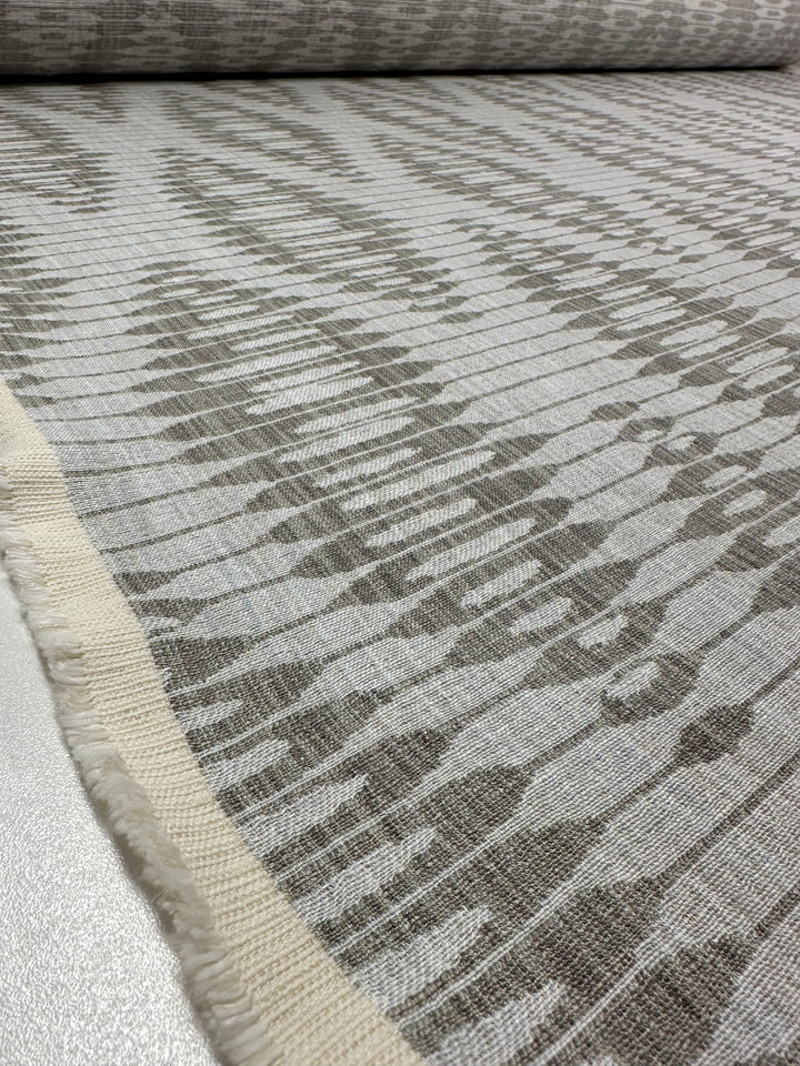 The Upholstery Jacquard - Ludwig Silver by Super Cheap Fabrics showcases an ikat-inspired gray and white abstract pattern with irregular, organic shapes. The jacquard weave adds depth, complemented by a rough border against the smooth 140cm fabric surface.