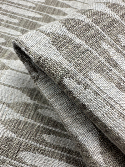 A close-up of Upholstery Jacquard - Ludwig Silver by Super Cheap Fabrics showcases the folded fabric's textured surface with a beige and gray geometric pattern, ikat-inspired appearance, visible fibers, and a slightly rustic look. Ideal for upholstery. Width: 140cm.