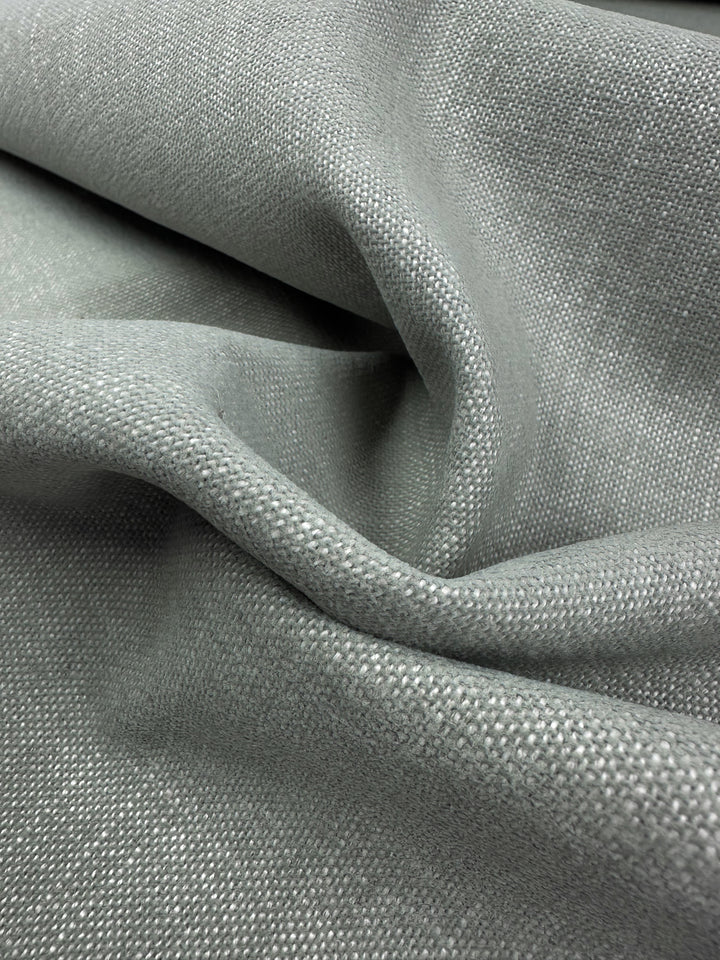 A close-up of Warm Silver upholstery fabric from Super Cheap Fabrics in a muted green shows its fine plain weave and lightweight, natural drape—perfect for adding subtle elegance to any room. Width: 142cm.