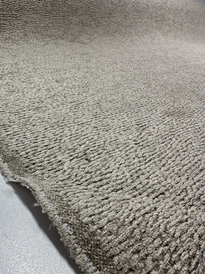 Close-up of a textured Upholstery Chenille Ezard fabric in Silver Taupe by Super Cheap Fabrics. The 145cm wide material has a velvety feel and an uneven surface, with its rolled edges against a gray background showcasing the fiber's subtle wave pattern.