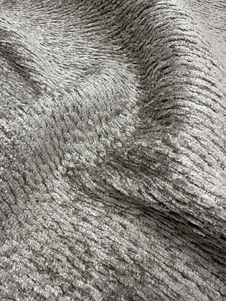 The Upholstery Chenille Ezard in Silver Taupe from Super Cheap Fabrics showcases a wavy, ribbed pattern with a silvery-gray color. It has an extra-heavy weight, velvety feel, and soft texture that shimmers elegantly under different lighting.