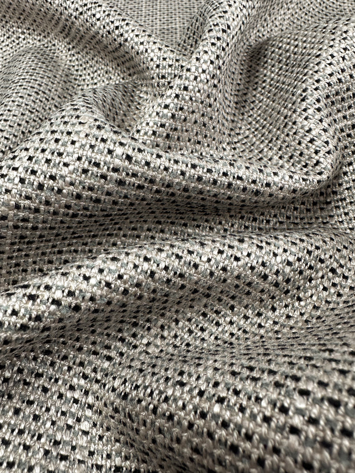 A close-up of Upholstery Logan - Rhino - 145cm, by Super Cheap Fabrics, reveals a heavy-weight fabric with a woven pattern in silver and black. The draped fabric forms soft folds and shadows that enhance its texture and luster.