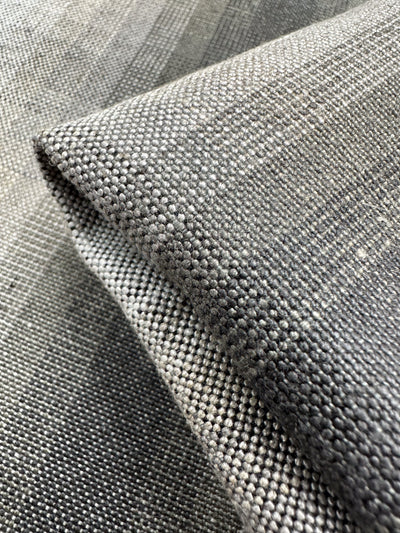 A close-up of Super Cheap Fabrics' Pure Linen - Natural - 148cm shows its textured gray fabric, featuring a woven pattern of small squares and shades ranging from light to dark. When folded, the fabric creates shadows and highlights that enhance its texture and depth.