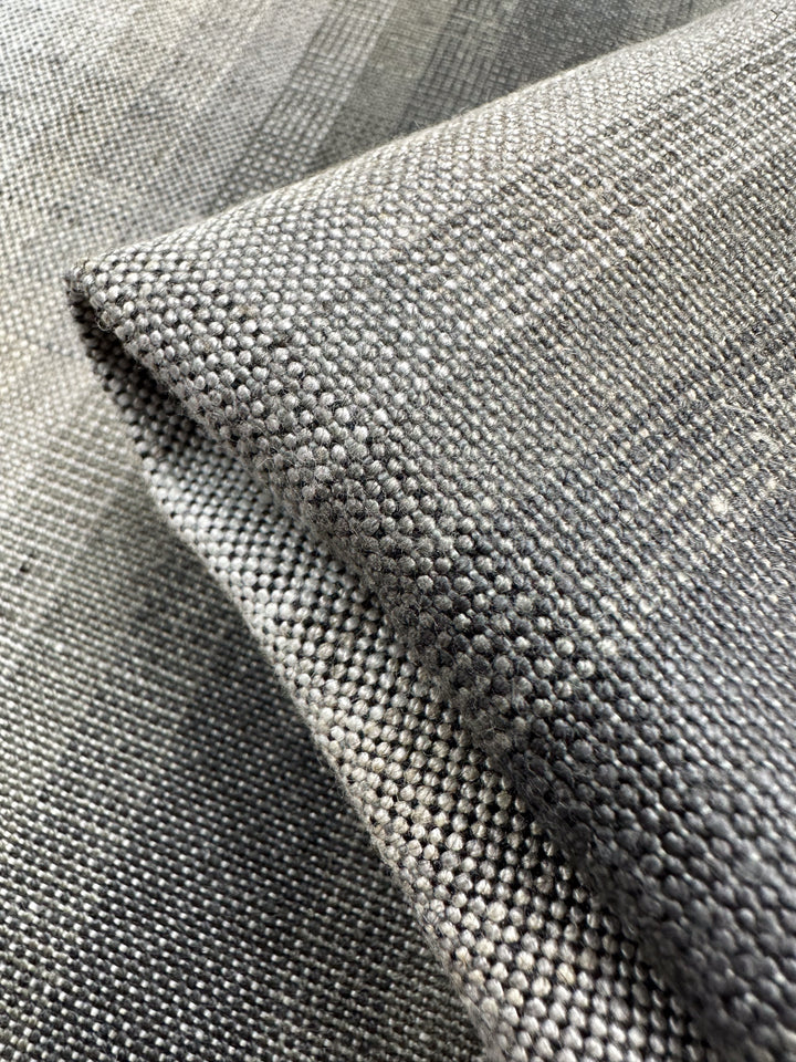 A close-up of Super Cheap Fabrics' Pure Linen - Natural - 148cm shows its textured gray fabric, featuring a woven pattern of small squares and shades ranging from light to dark. When folded, the fabric creates shadows and highlights that enhance its texture and depth.