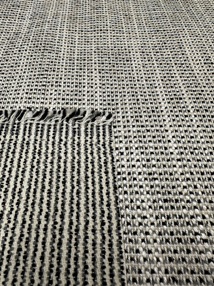 The Upholstery Logan - Salt & Pepper by Super Cheap Fabrics features a gray woven textile with a small fringed edge, checkered pattern in varying gray shades, and an open-weave grid design. Its textured appearance is enhanced by the tight weave and slightly irregular fringe.